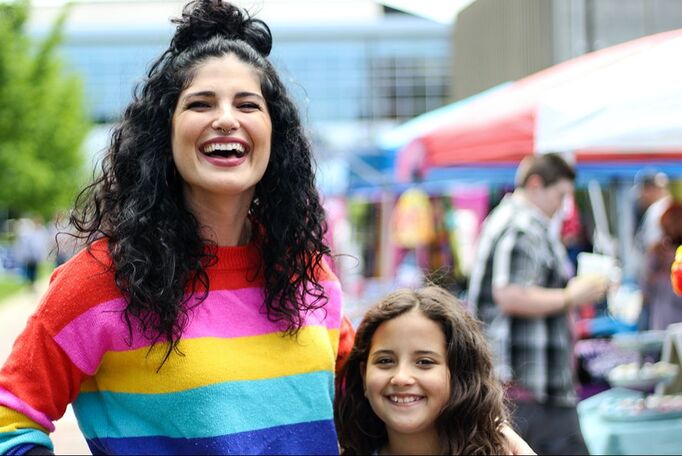 A woman in a striped rainbow sweater stands with her arm around a young girl at a Pride festival.